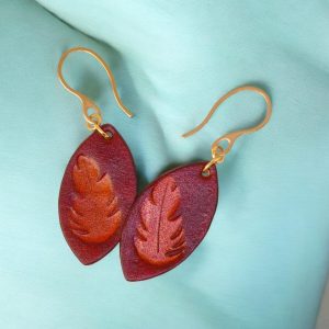 Boucles d'oreilles forme poisson en cuir teinture cognac, estampage fougère, vues de face se chevauchant, suspendues à des boucles dorées sur fond dégradé