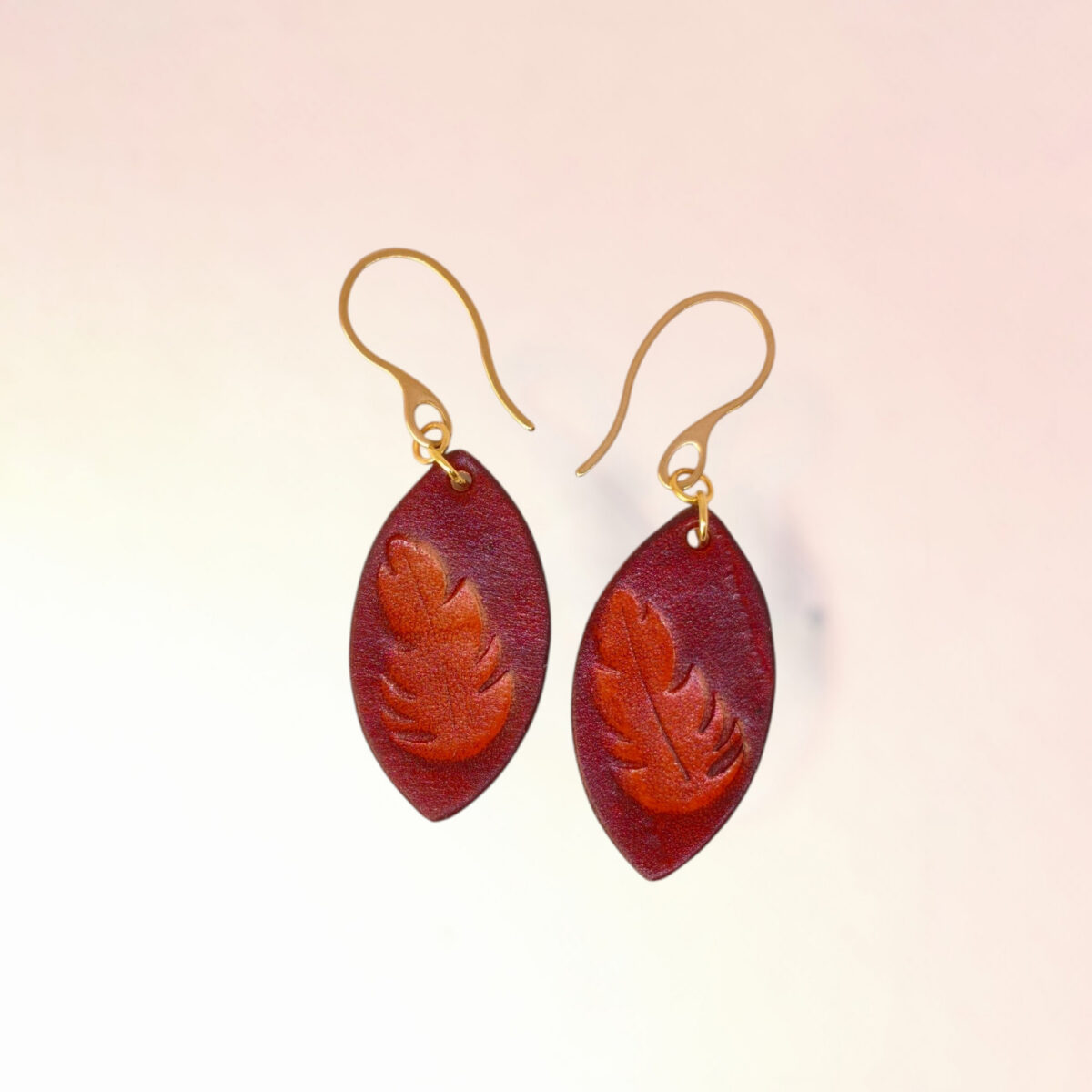 Boucles d'oreilles forme poisson en cuir teinture cognac, estampage fougère, vues de face, suspendues à des boucles dorées sur fond dégradé