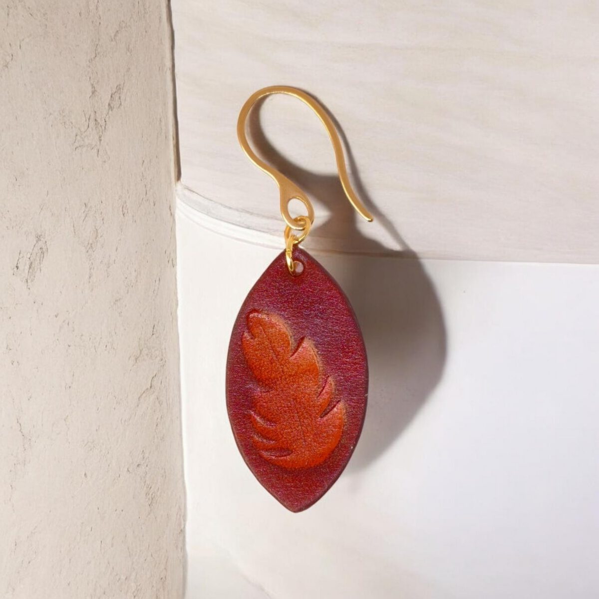 Boucle d'oreille forme poisson en cuir teinture cognac, estampage fougère, vue de face, suspendue à une boucle dorée, fond beige