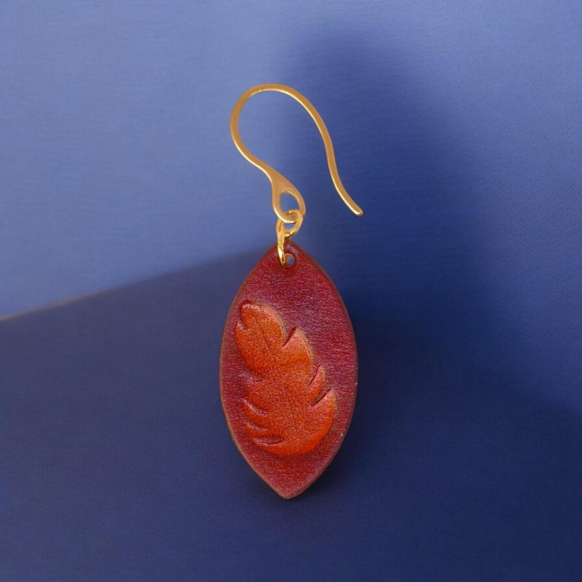 Boucle d'oreille forme poisson en cuir teinture cognac, estampage fougère, vue de face, suspendue à une boucle dorée, fond design