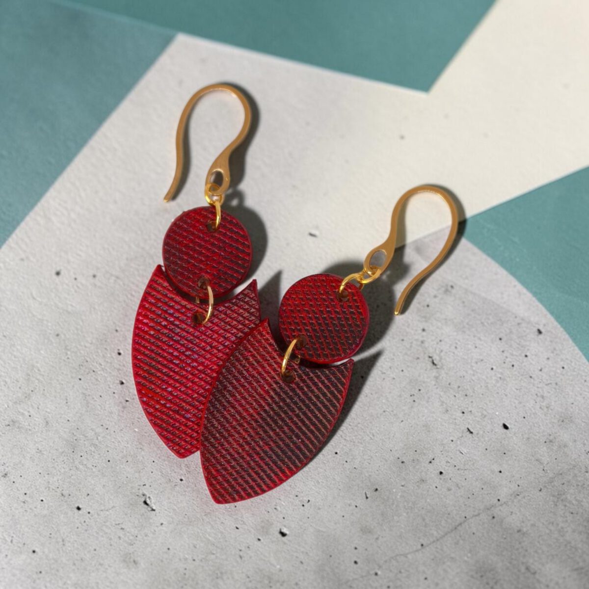 Boucles d'oreilles forme double rond + écu en cuir teinture rouge, texturage lime fine et finition vieillie, vues de face se chevauchant, suspendues à des boucles dorées sur béton