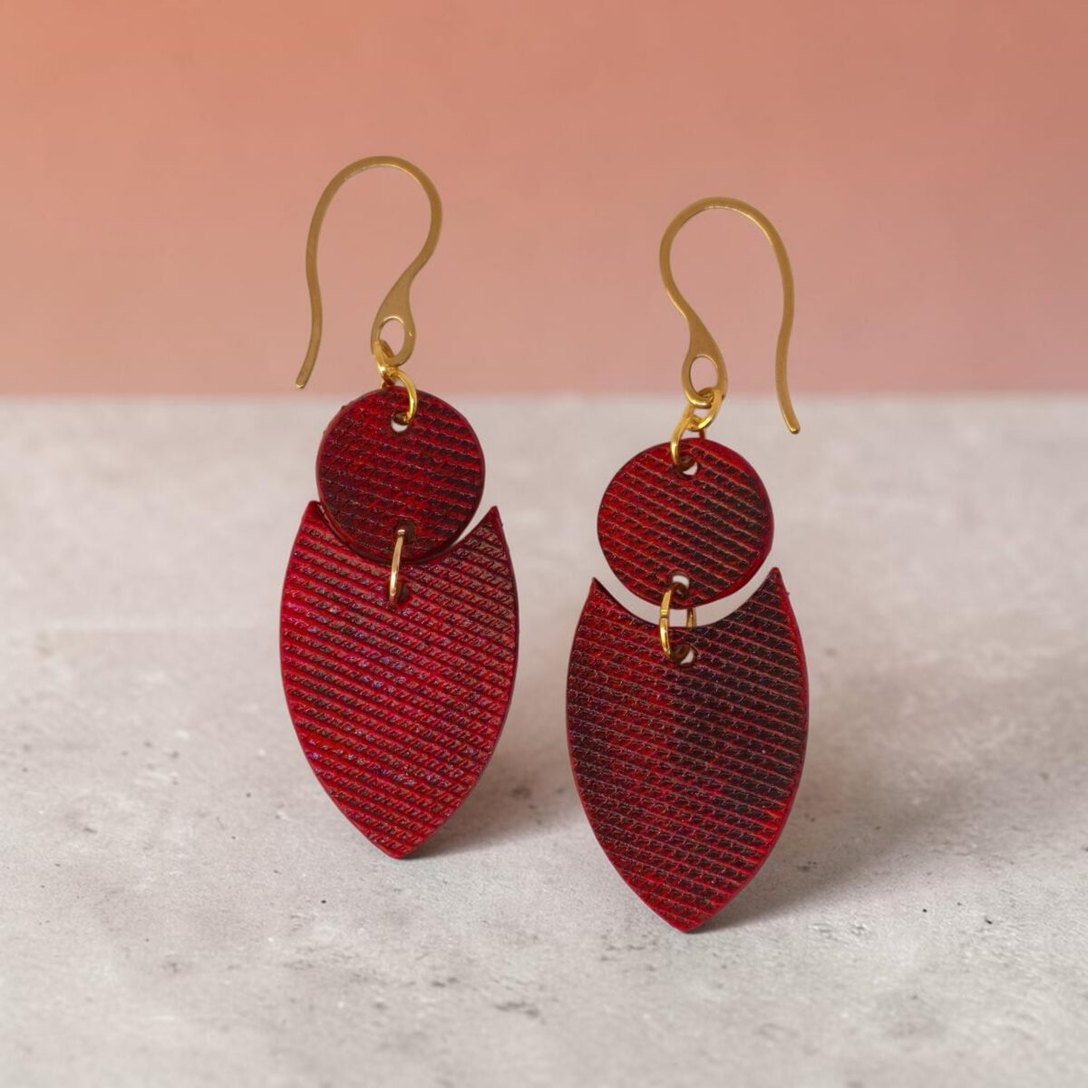 Boucles d'oreilles forme double rond + écu en cuir teinture rouge, texturage lime fine et finition vieillie, vues de face, suspendues à des boucles dorées sur béton