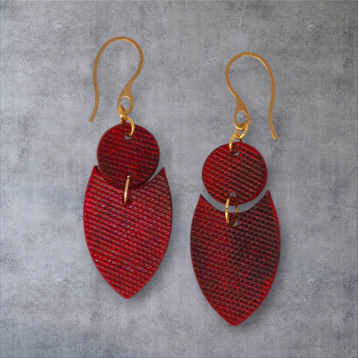 Boucles d'oreilles forme double rond + écu en cuir teinture rouge, texturage lime fine et finition vieillie, vues de face, suspendues à des boucles dorées sur fond gris
