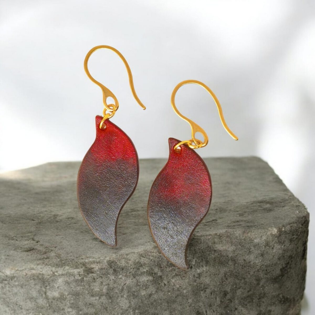Boucles d'oreilles en cuir teintées en noir et rouge, vues de face, suspendues à des crochets dorés sur pierre