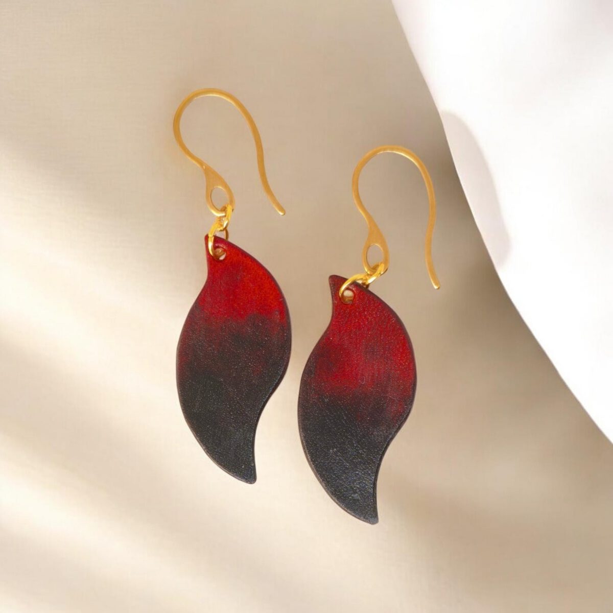 Boucles d'oreilles en cuir teintées en noir et rouge, vues de face, suspendues à des crochets dorés sur fond design