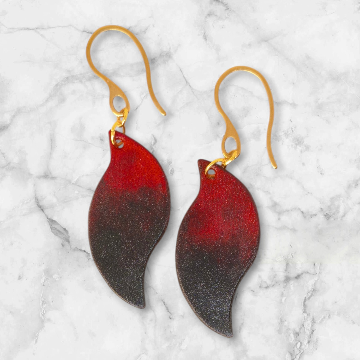 Boucles d'oreilles en cuir teintées en noir et rouge, vues de face, suspendues à des crochets dorés posées sur marbre