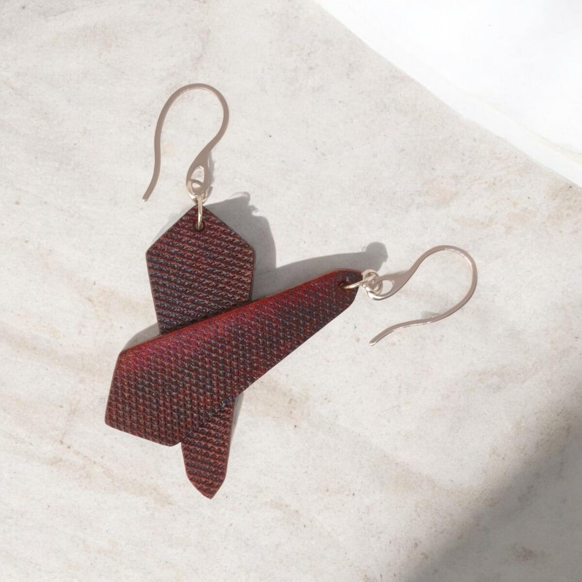 Boucles d'oreilles asymétriques en cuir teinture acajou, vues de face se chevauchant, suspendues à des crochets argentés sur fond design