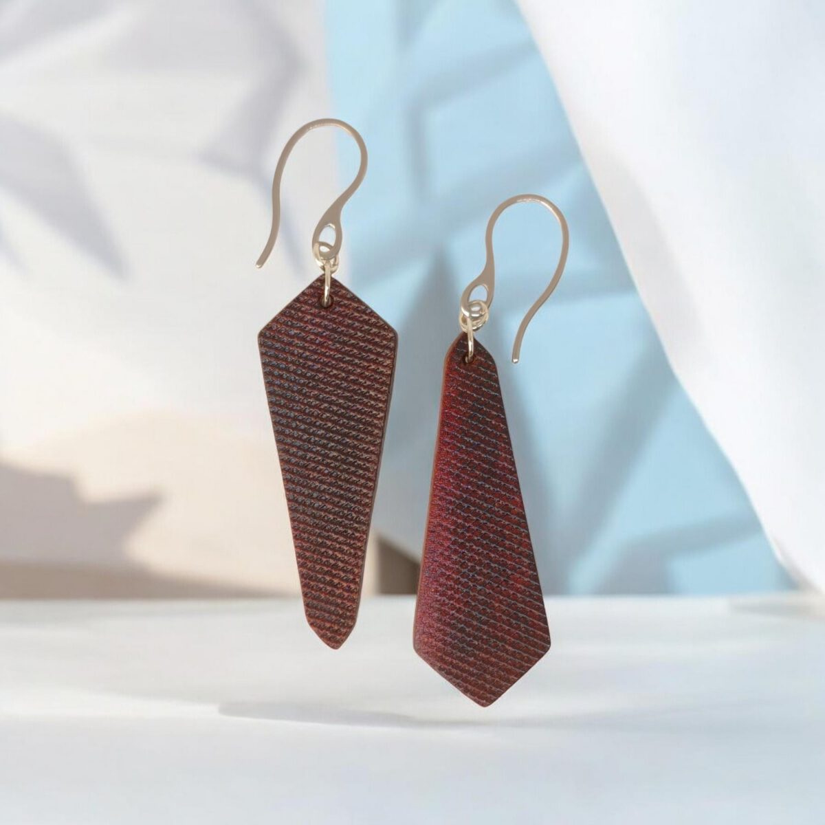 Boucles d'oreilles asymétriques en cuir teinture acajou, vues de face, suspendues à des crochets argentés sur fond design