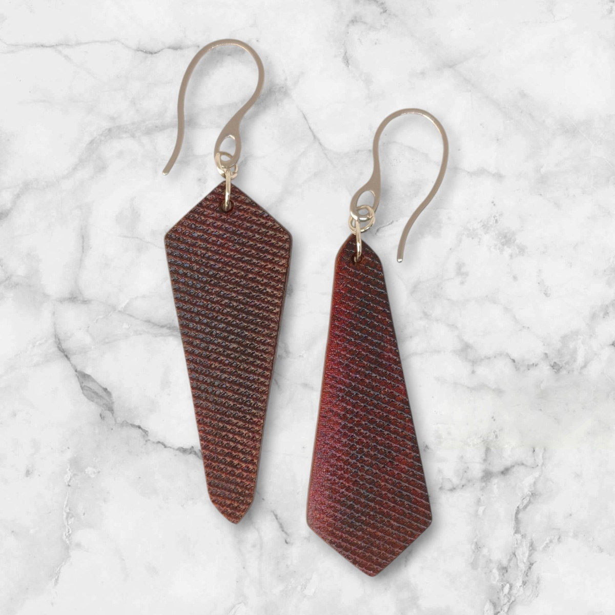 Boucles d'oreilles asymétriques en cuir teinture acajou, vues de face, suspendues à des crochets argentés sur fond marbré