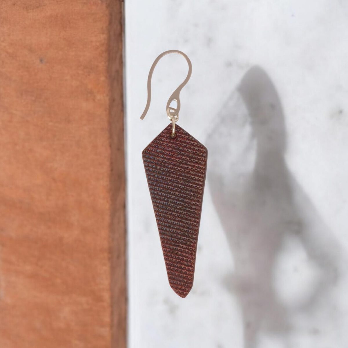 Boucle d'oreille en cuir, teinture acajou et finition lime vieillie, vue de face, suspendue à un crochet argenté sur fond design