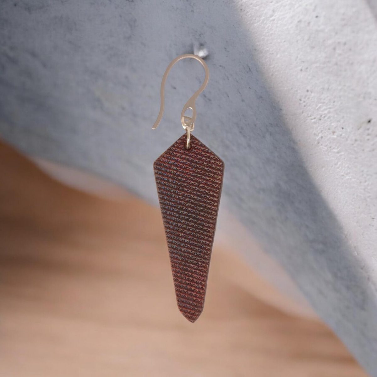 Boucle d'oreille en cuir, teinture acajou, vue de face, suspendue à un crochet argenté, fond design