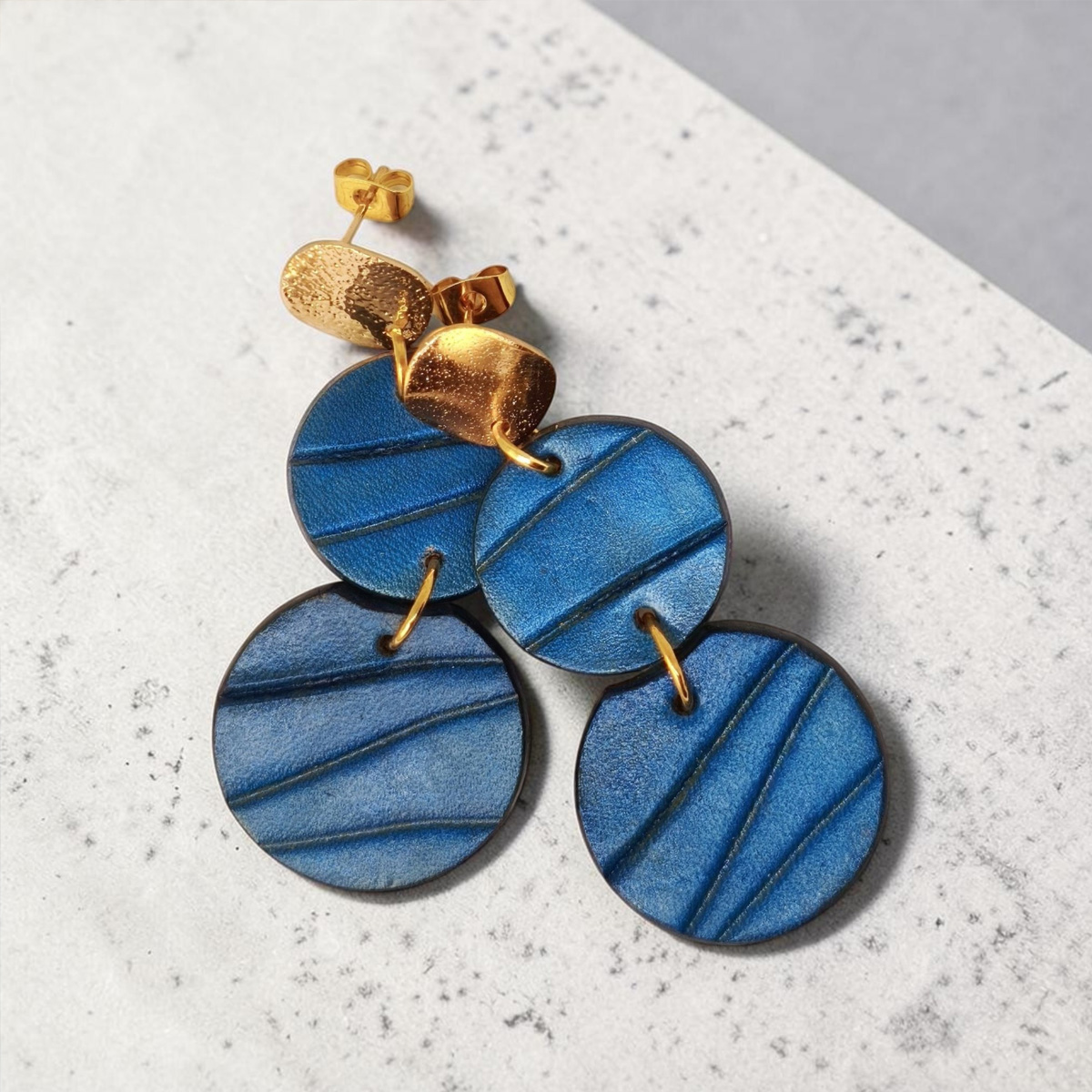 Boucles d'oreilles rondes doubles en cuir teinture bleue, finition galet, vues de face se chevauchant, suspendues à des boucles dorées sur béton