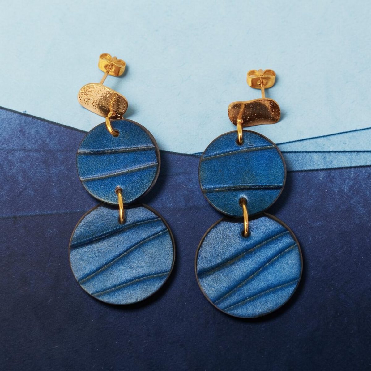 Boucles d'oreilles rondes doubles en cuir teinture bleue, finition galet, vues de face, suspendues à des boucles dorées, fond design