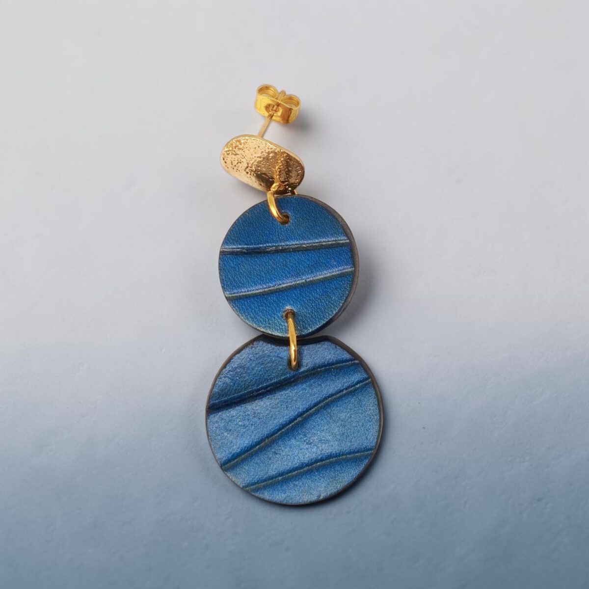 Boucle d'oreille rondes doubles en cuir teinture bleue, finition galet, vue de face, suspendue à une boucle dorée, fond dégradé