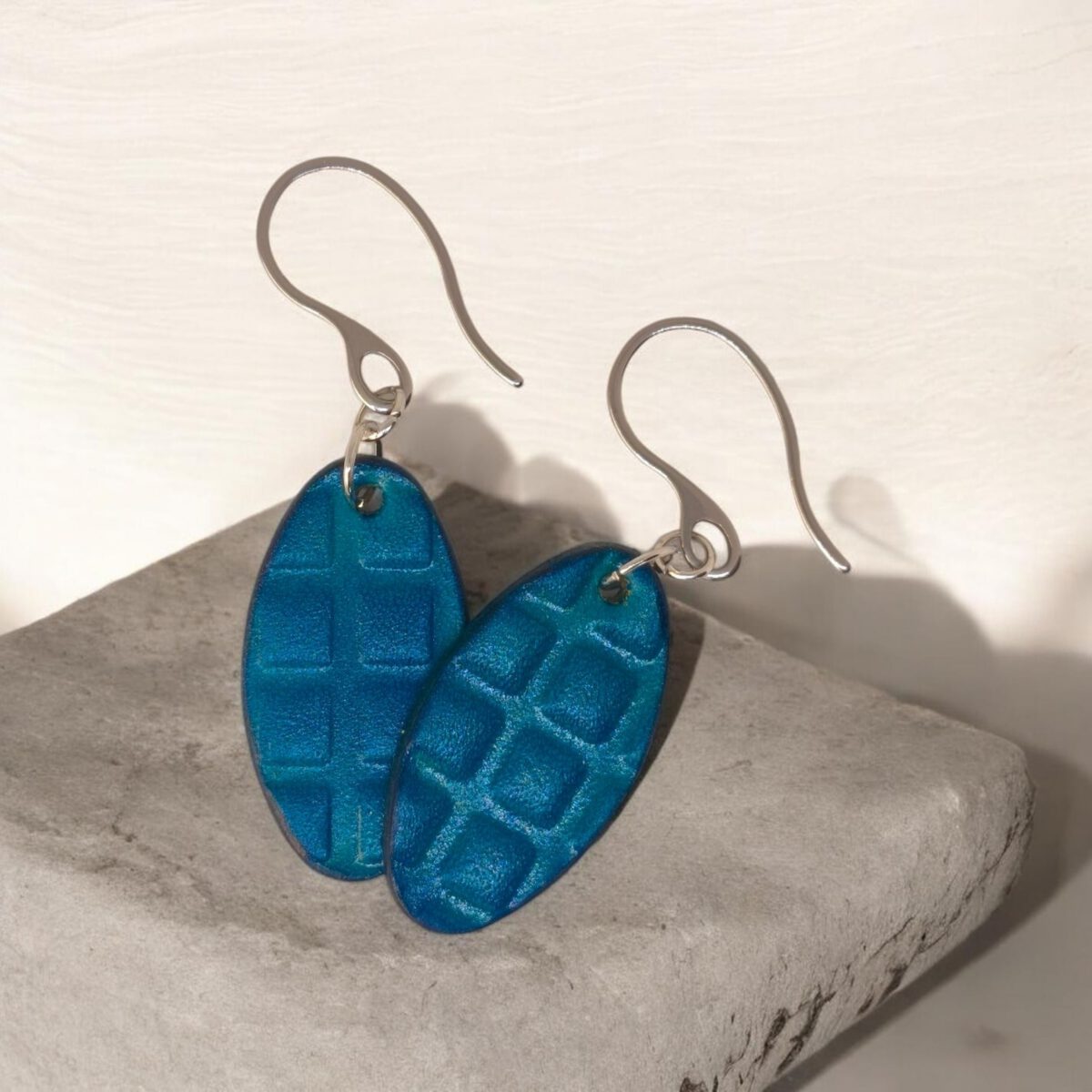 Boucles d'oreilles ovales en cuir teinte bleue, texturage carré, vues de face se chevauchant, suspendues à des boucles argentées sur pierre