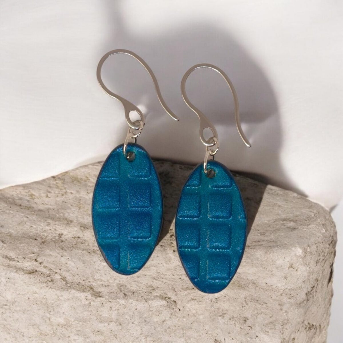Boucles d'oreilles ovales en cuir teinte bleue, texturage carré, vues de face, suspendues à des boucles argentées sur pierre