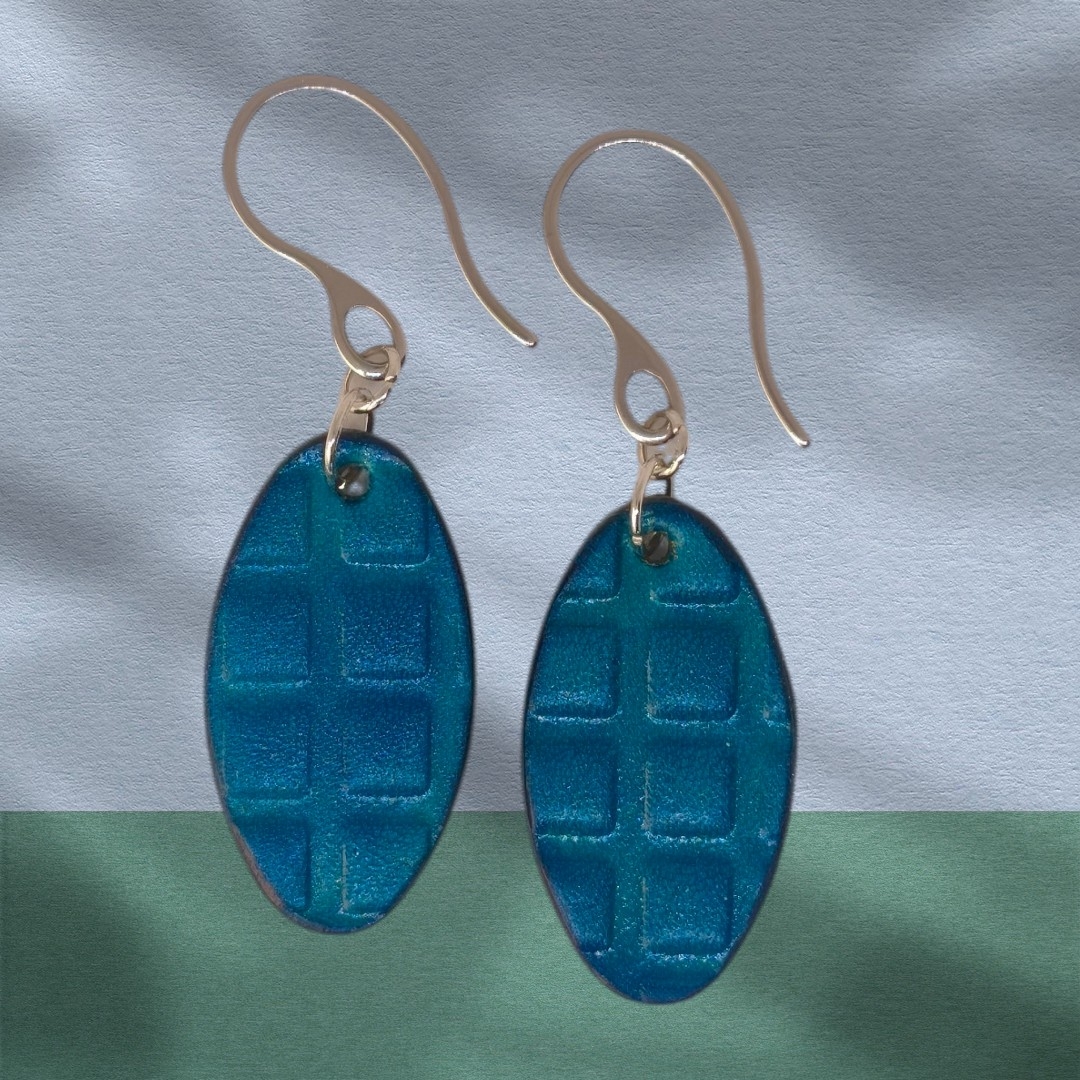 Boucles d'oreilles ovales en cuir teinte bleue, texturage carré, vues de face, suspendues à des boucles argentées sur fond design