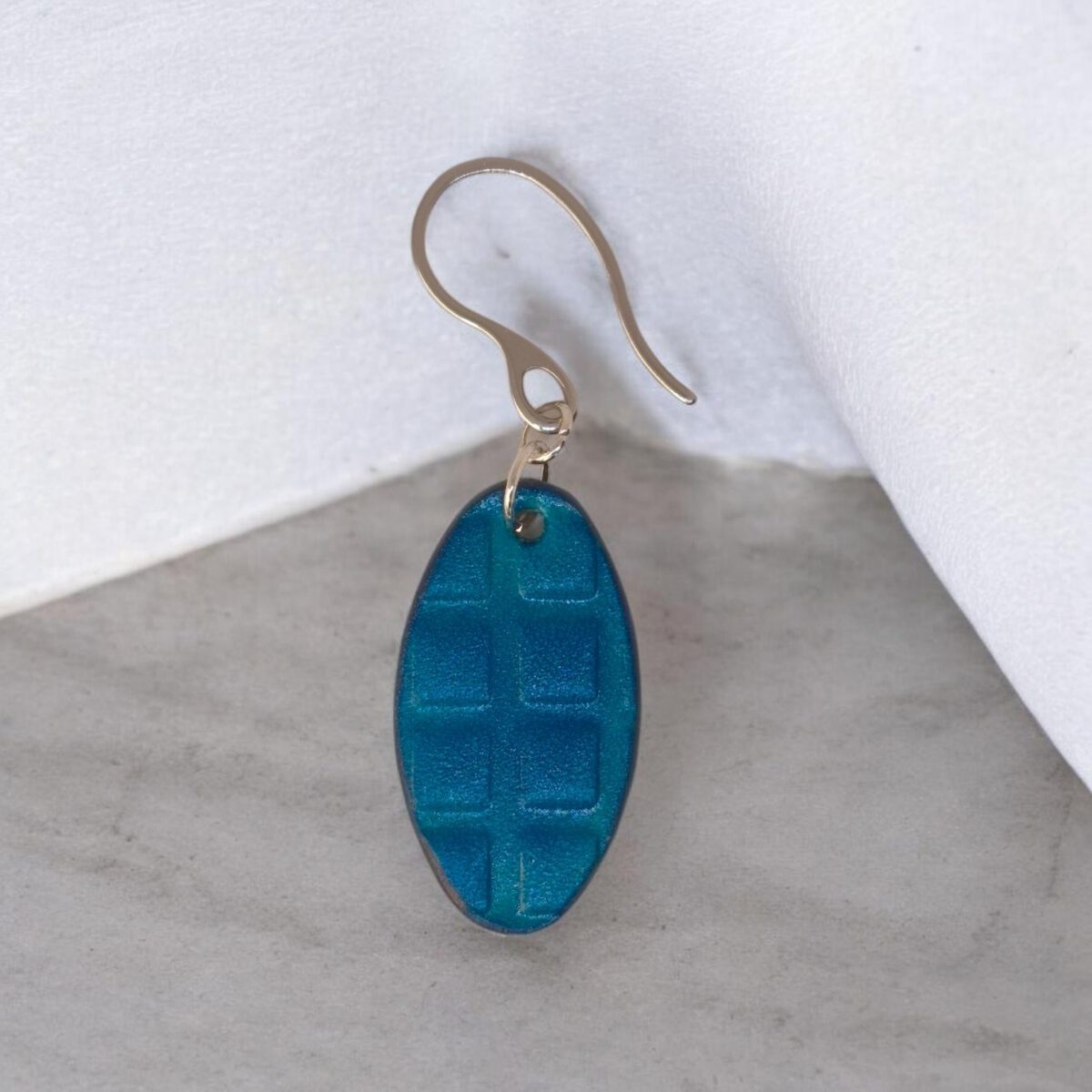 Boucle d'oreille ovale en cuir teinte bleue, texturage carré, vue de face, suspendue à une boucle argentée, fond gris