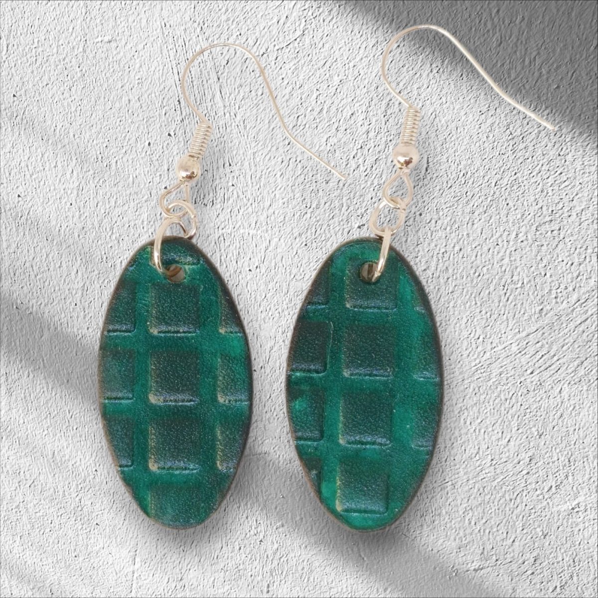 Boucles d'oreilles ovales en cuir teinte vert d'eau, texturage carrés, vues de face, suspendues à des boucles argentées sur crépi