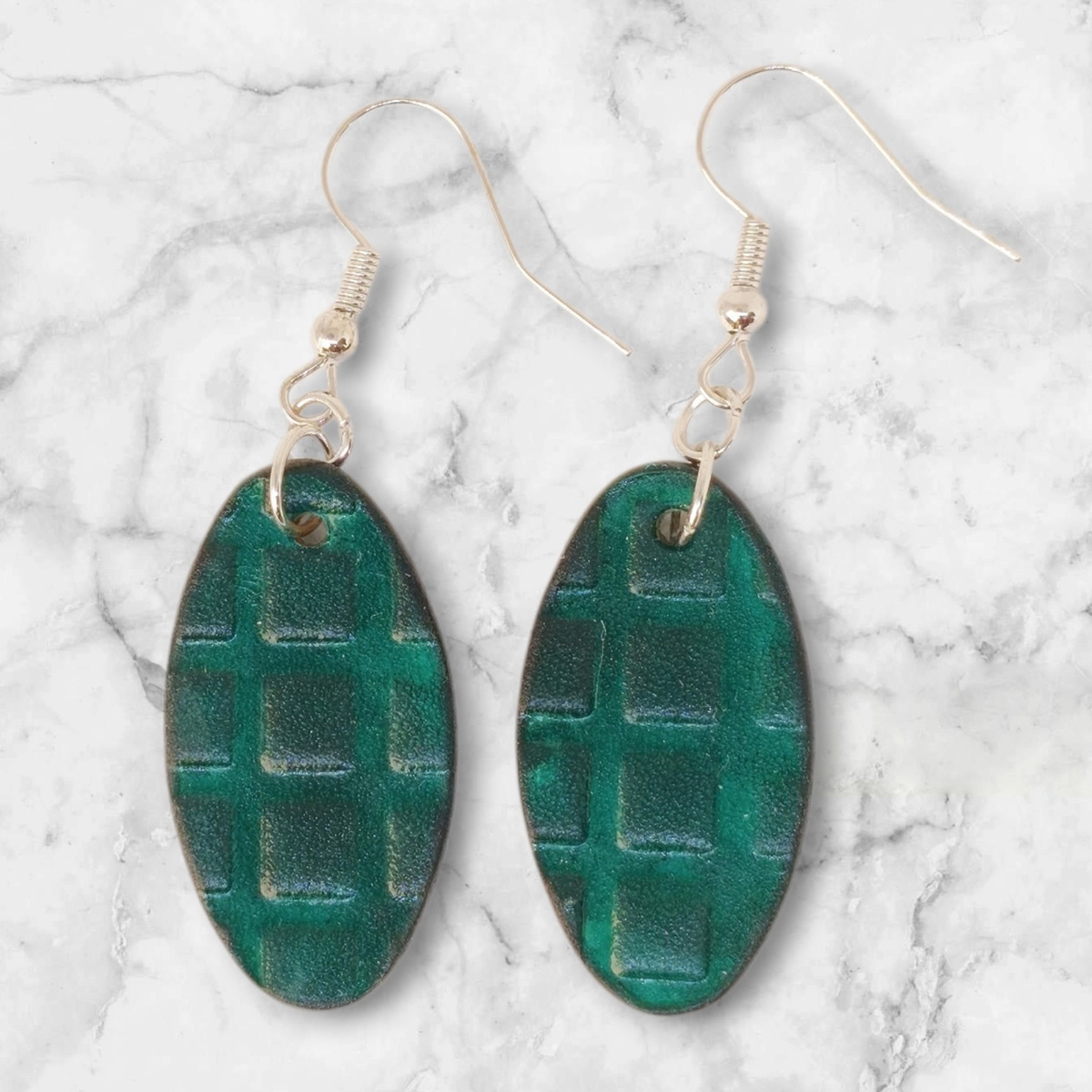 Boucles d'oreilles ovales en cuir teinte vert d'eau, texturage carrés, vues de face, suspendues à des boucles argentées sur marbre