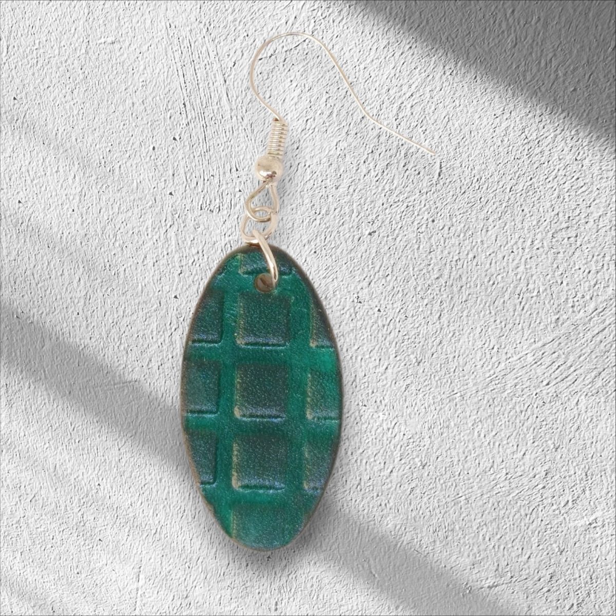 Boucle d'oreille ovale en cuir teinte vert d'eau, texturage carrés, vue de face, suspendue à une boucle argentée, fond crépi