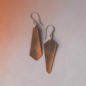Boucles d'oreilles asymétriques en cuir, huilées teinte naturelle, vues de face, suspendues à des crochets argentés sur fond dégradé