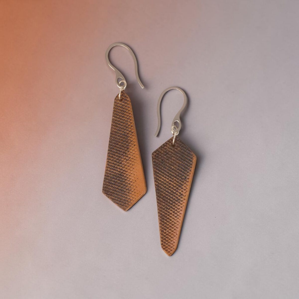Boucles d'oreilles asymétriques en cuir, huilées teinte naturelle, vues de face, suspendues à des crochets argentés sur fond dégradé