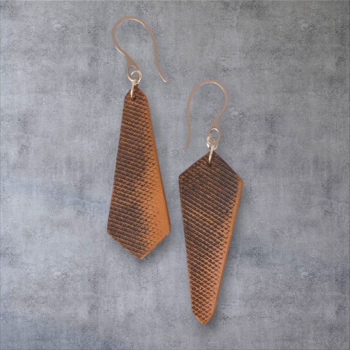 Boucles d'oreilles asymétriques en cuir, huilées teinte naturelle, vues de face, suspendues à des crochets argentés sur fond gris