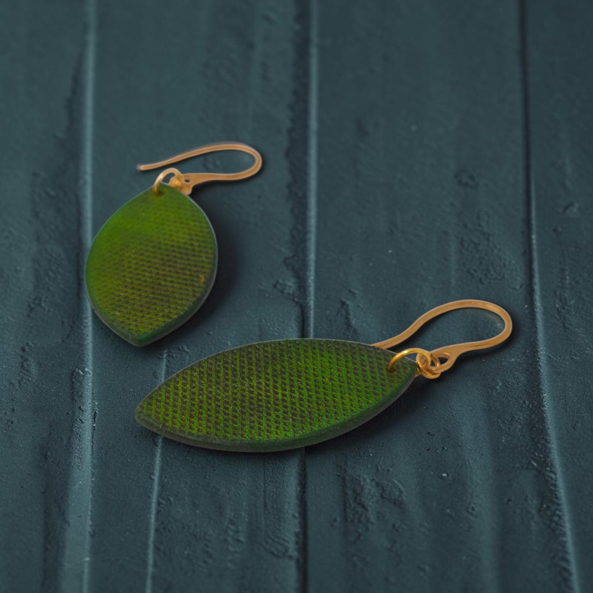 Boucles d'oreilles forme poisson en cuir teinture vert forêt, texturage lime épaisse, vue latérale, suspendues à des boucles dorées fond design