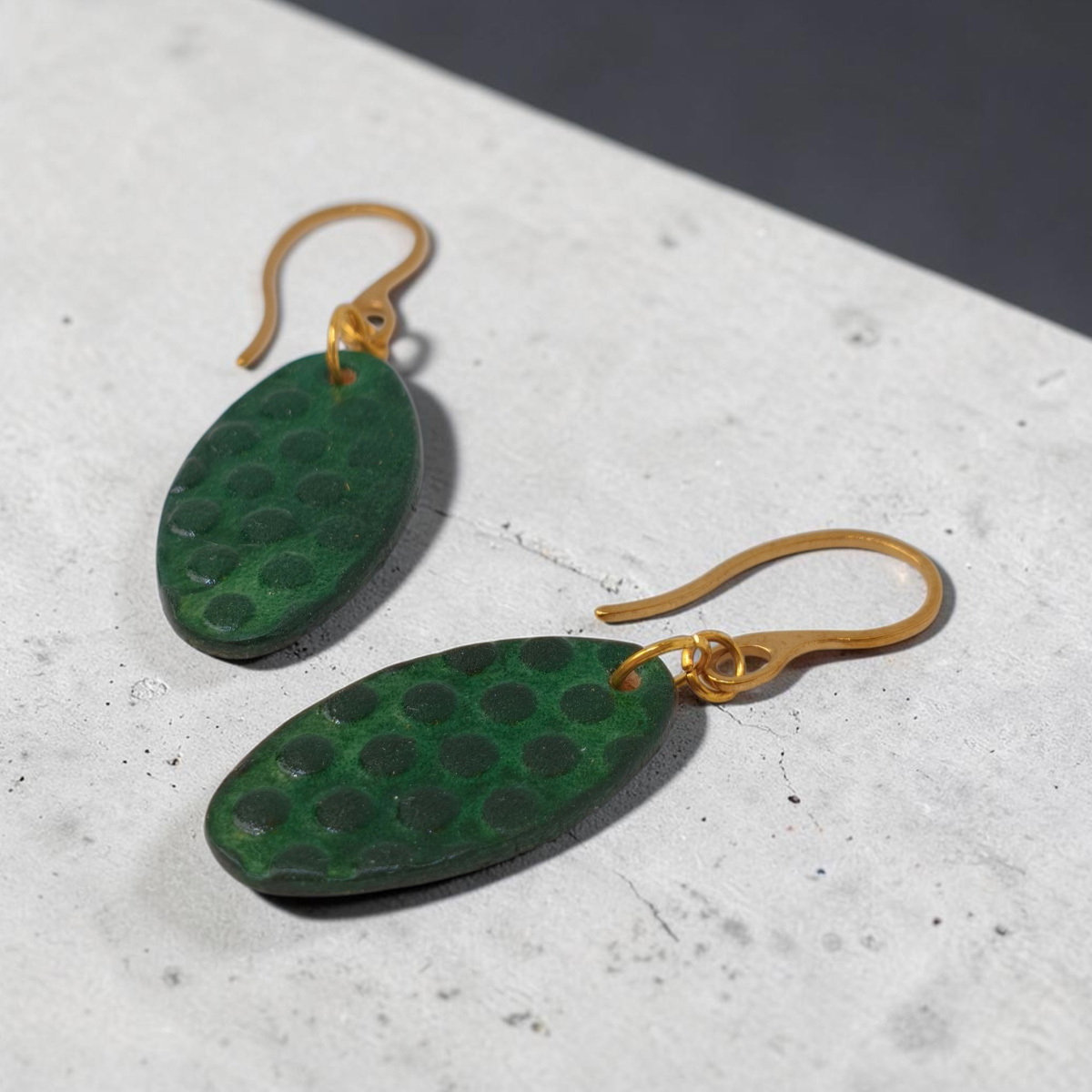 Boucles d'oreilles ovales en cuir teinte vert forêt, texturage ronds, vue latérale, suspendues à des boucles dorées sur fond gris