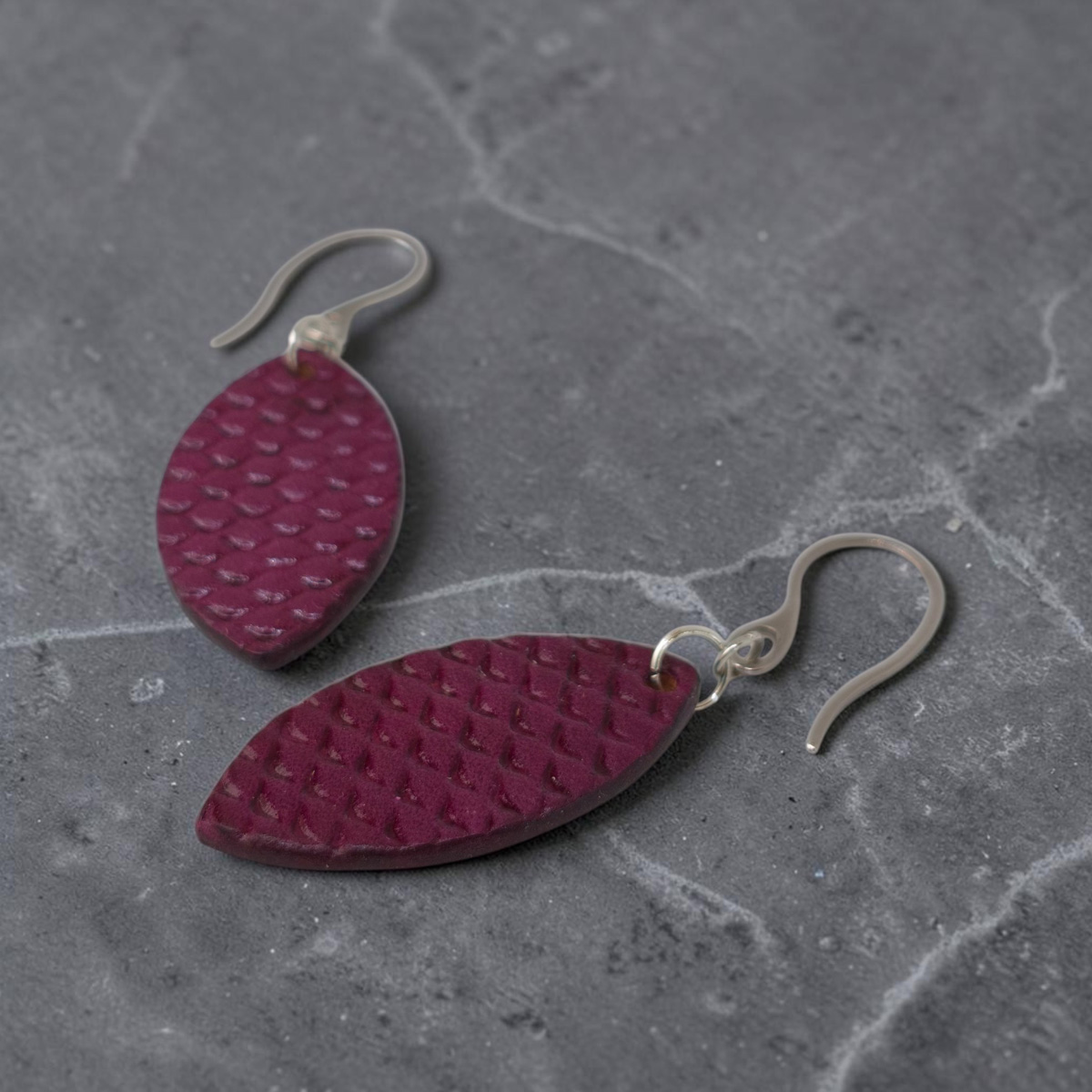 Boucles d'oreilles forme poisson en cuir teinture violet, texturage poisson fin, vue latérale, suspendues à des boucles argentées sur sable