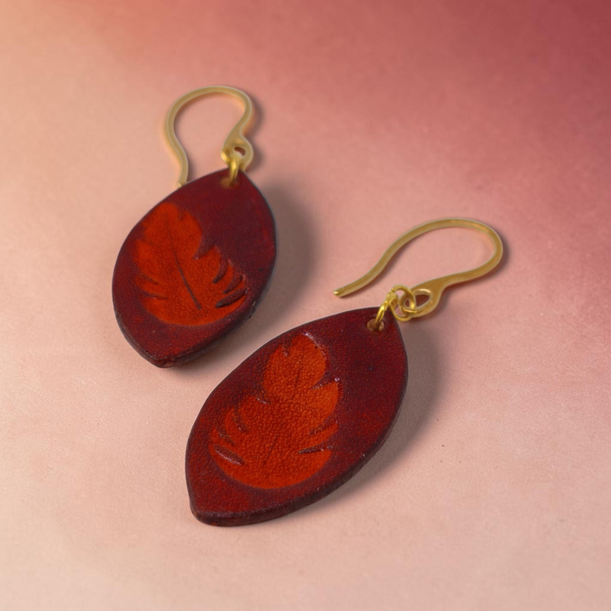 Boucles d'oreilles forme poisson en cuir teinture cognac, estampage fougère, vue latérale, suspendues à des boucles dorées sur marbre